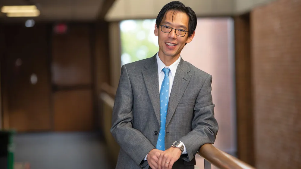 A man of Asian descent smiles as poses for this photo wearing a suit. He casually leans on a railing inside a university building. He looks like the kind of professor you'd want to take a class from--good natured and intelligent.