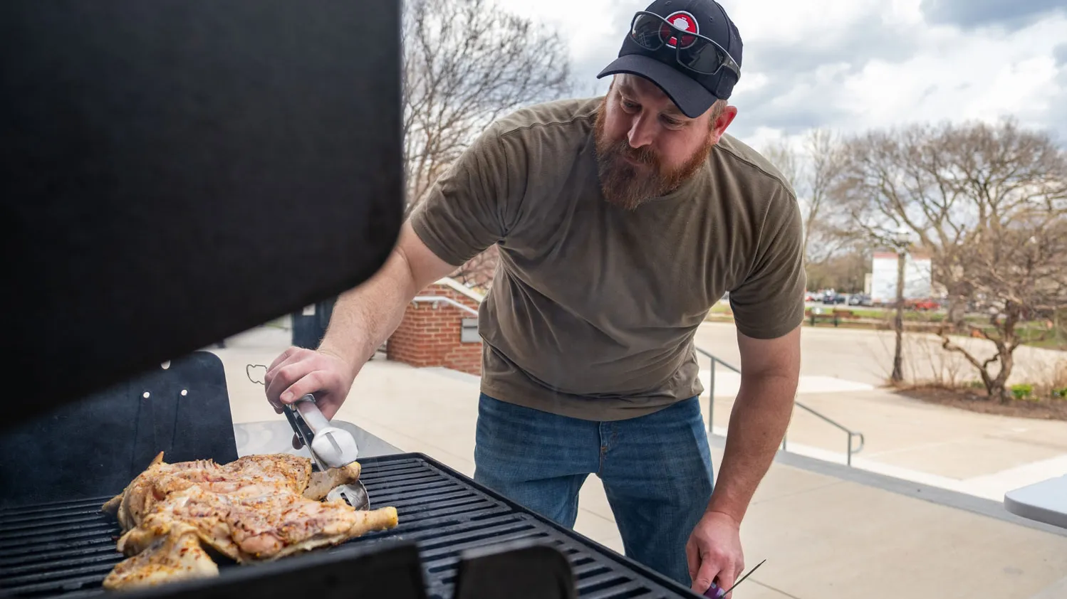 Michael Cressman leans over a grill while cooking a chicken. Cressman is a white man wearing a T-shirt, jeans and ballcap. He looks focused as he uses tongs to check the underside on Ohio State’s campus.