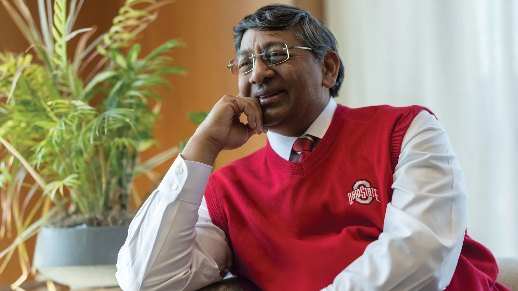Ravi Bellamkonda leans one elbow on the arm of a couch as he leans into it and chuckles. He wears a scarlet Ohio State sweater vest, glasses and has salt-and-pepper tussled hair. He appears to be invested in a conversation