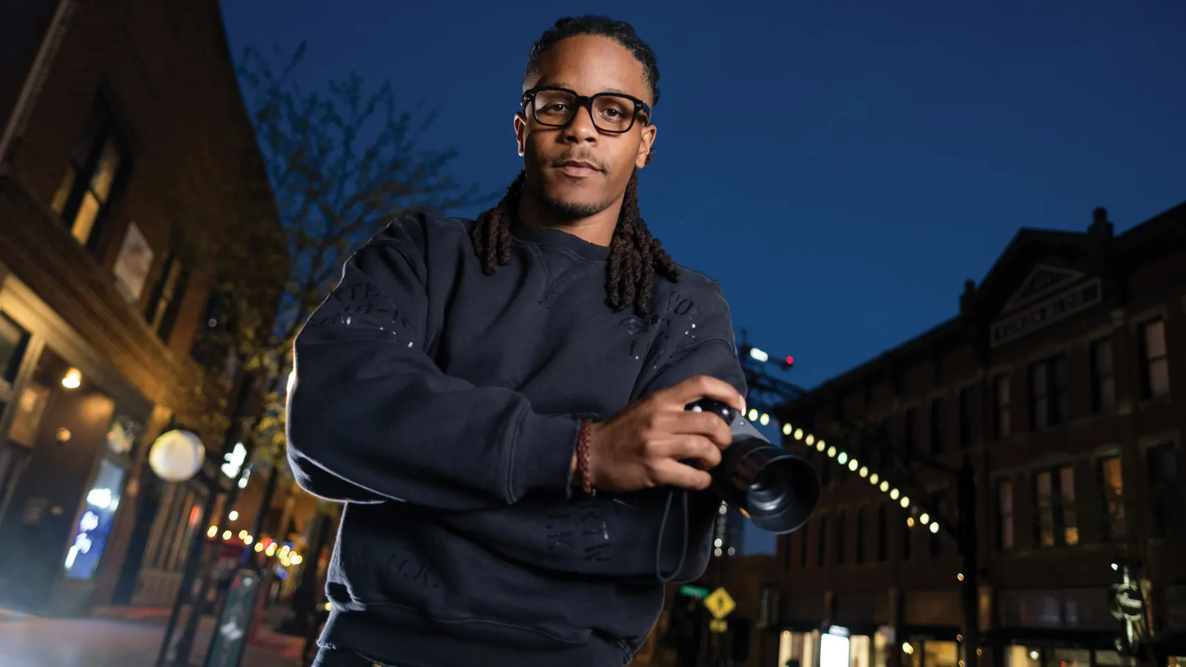 Justen Turner is a young black man wearing thick-framed glasses and a sweatshirt. He looks focused as he poses for this portrait holding a small video camera. His hair is dreads pulled back in a low ponytail. He looks like he's competent, professional and thoughtful—or like someone you'd trust to make a movie about yourself or a loved one. He's photographed in the Short North after the sun has set.
