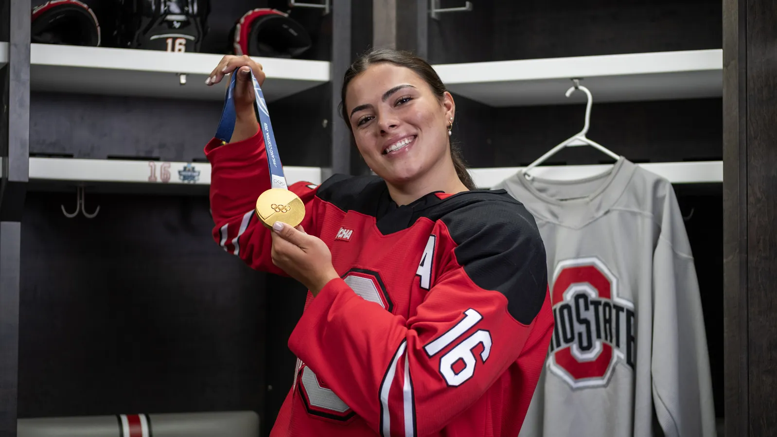 Joy Dunne smiles as she proudly shows off her gold medal in the locker room for Ohio State women's hockey. She wears her team uniform. 