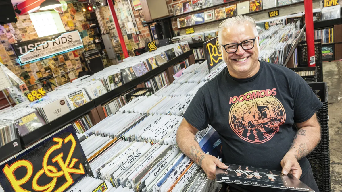 Used Kids owner Greg Hall laughs as he is photographed in the record store, wearing thick-framed glasses and an artful T-shirt for the hard rock band 1000 Mods.