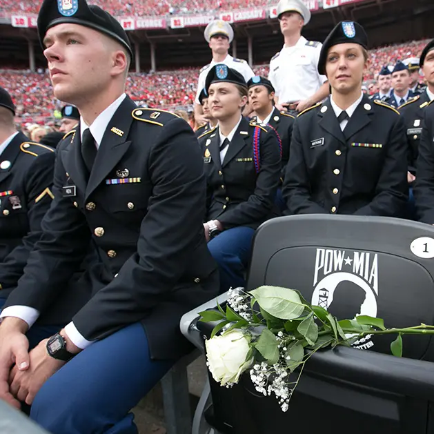 Military men and women in uniform sitting in Ohio Stadium. There is an empty chair with white roses in the seat in honor of all POW-MIA.
