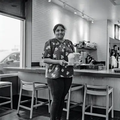 A woman of Indian or Middle Eastern descent carries a cup holder with drinks that might be coffees and/or milk shakes. She has the kind of smile in which her lips actually turn down as her dimples show. She’s in a coffee shop.