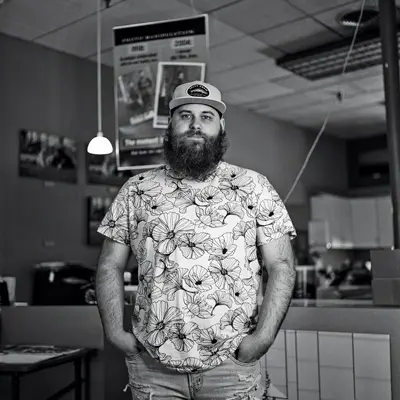 A younger white man with a bushy dark beard, ballcap and a flowered T-shirt stands with his hands tucked in his pockets inside a diner.
