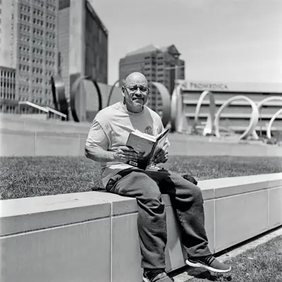 An older bald man holds open a hardback book as he sits on a low wall in a downtown of an unrecognizable city. His expression is serious; whether from the bright sunlight shining in his eyes or the subject matter of his book, it’s hard to tell. It’s hard to discern his race.