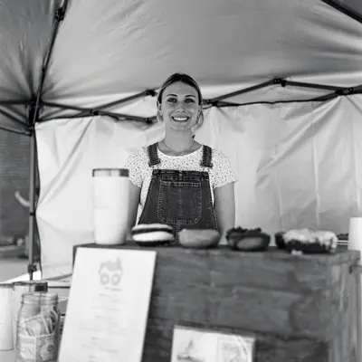 A young white woman wearing bib overalls stands proudly behind a makeshift counter topped with specialty donuts in a tent that looks to be at an outdoor farmers market. Her hair is pulled back in a ponytail and she has a big smile.