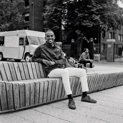 A younger black man wearing business casual clothes and a zip-up jacket sits confidently on a modern-looking bench in a neighborhood. His backpack is next to him. He has short dreadlocks pulled back at the crown of his head and a big smile.