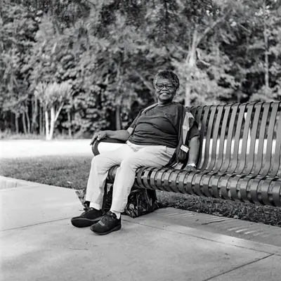 An older black woman offers a friendly smile as she rests on a bench in a city park. She has big-lense glasses, short hair that is graying and a trash bag under her seat, as if she’s been picking up trash.