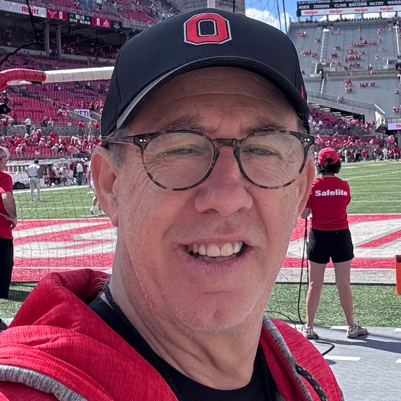 Mike Larsen smiles while on the sideline of Ohio Stadium. He wears an Ohio State hat and sweatshirt, as well as round glasses.