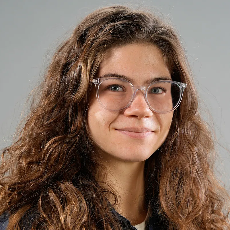 Mattie Mueller softly smiles as poses for a portrait. She has long curly hair and clear-rimmed glasses