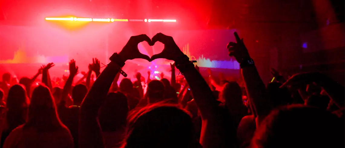 Person making a heart with their hands in a crowd of people dancing by a stage with red lights and fog