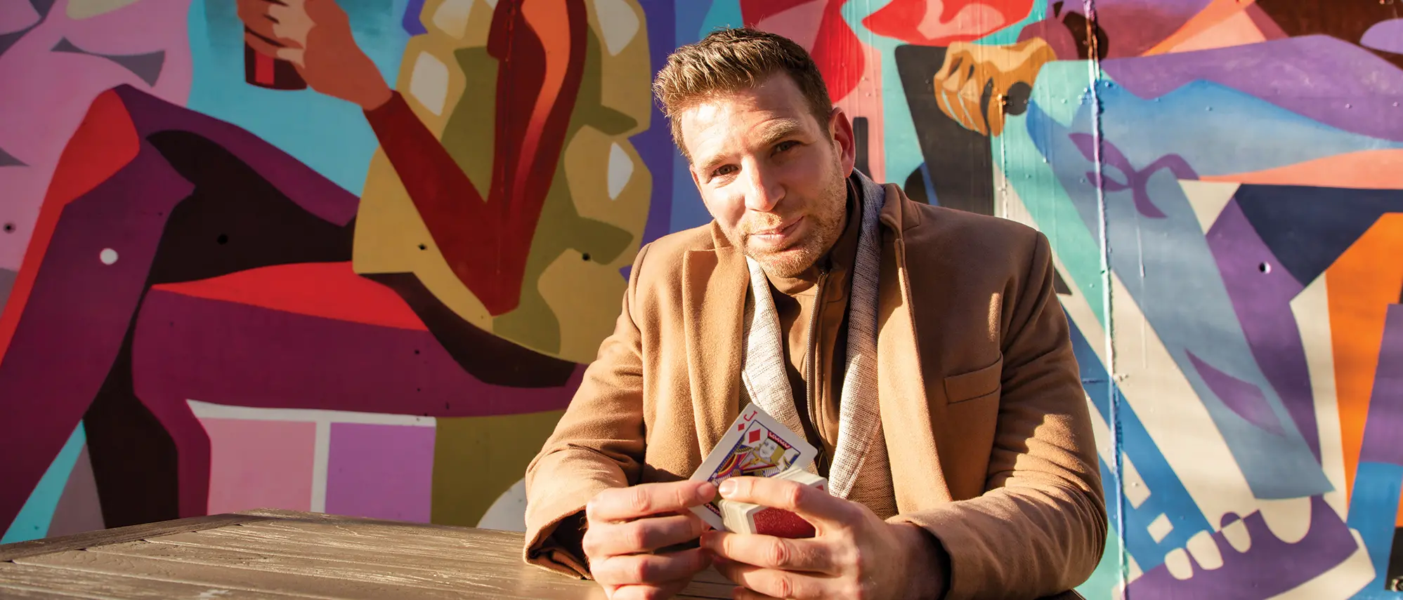 Smiling with his eyes, Joshua Jay holds a jack of diamonds while sitting at an outside mural
