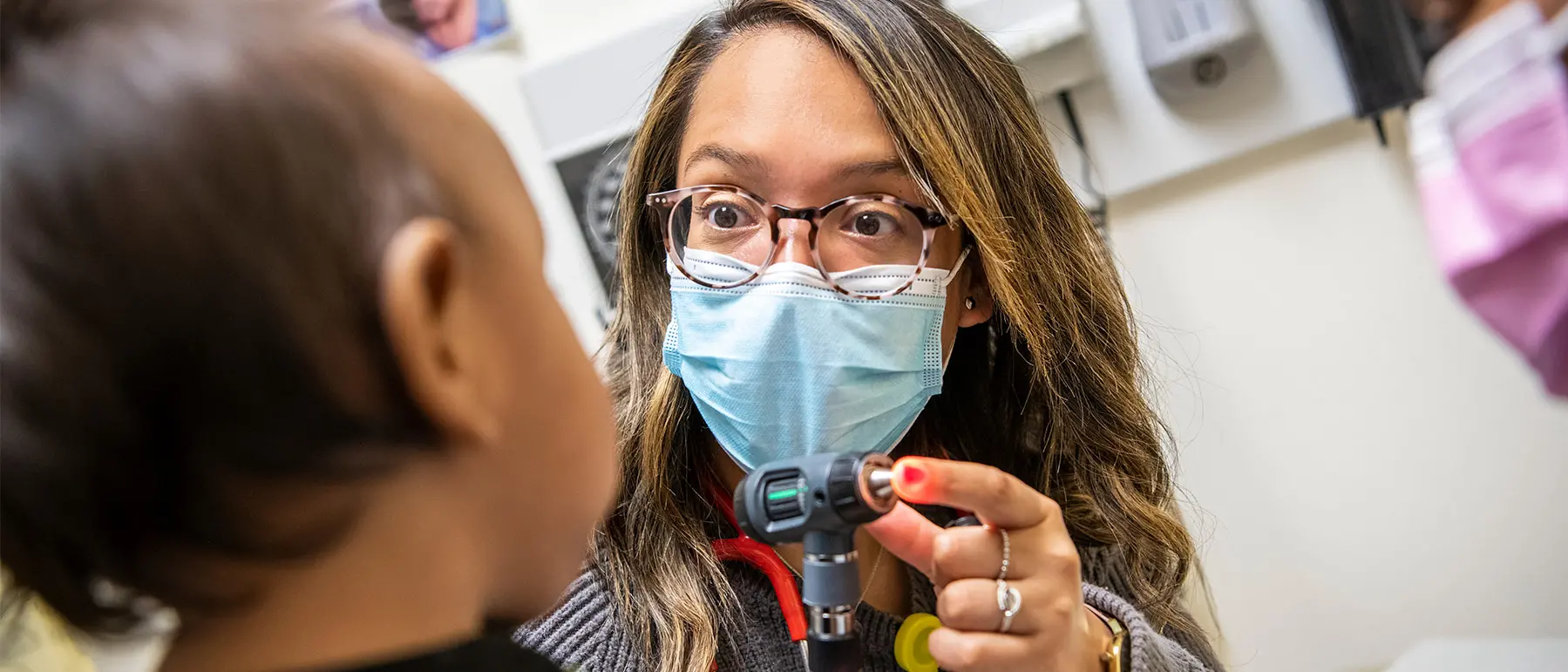 A young doctor of Guatemalan descent wears round tortoiseshell glasses, a surgical mask and a wide-eyed expression as she connects with a toddler. She’s showing the little girl the tool she’ll use to look in her ears.