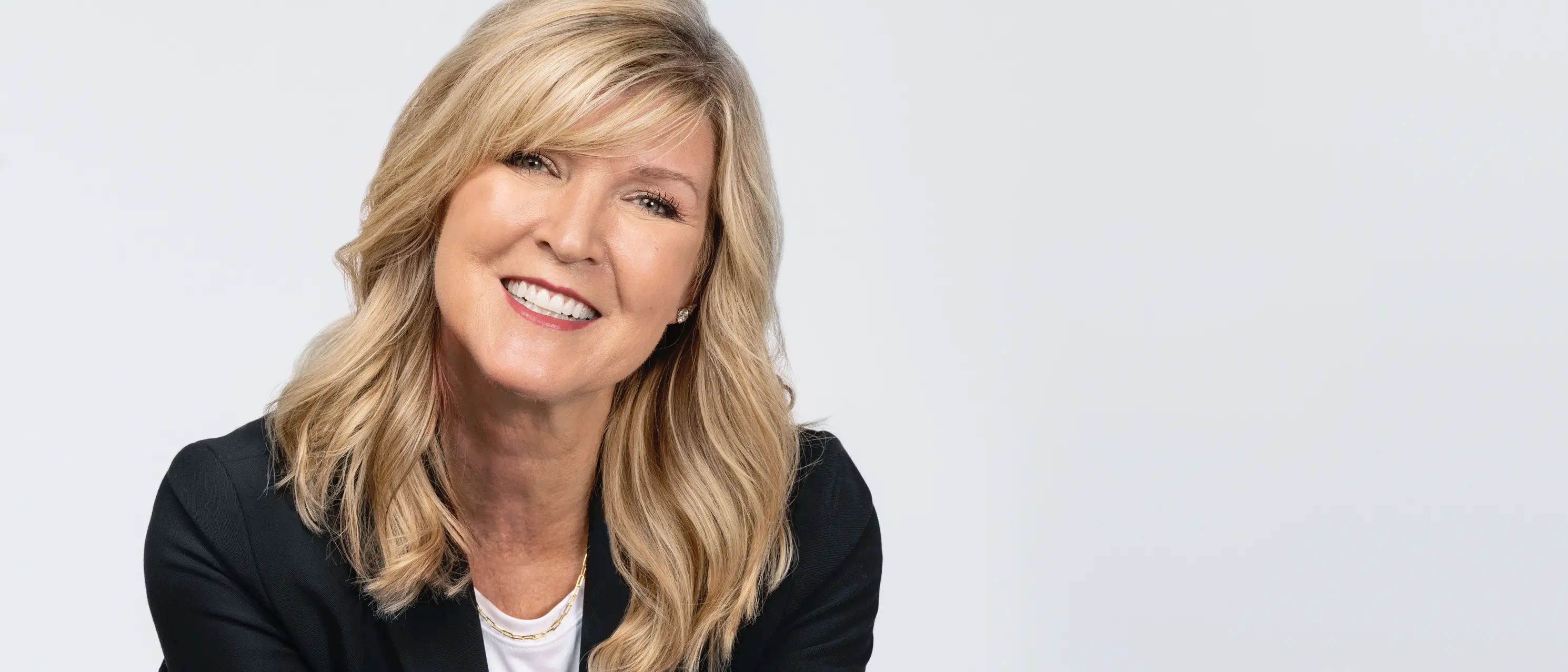 A white woman wears a pepperoni pizza T-shirt, under a black suit jacket, with black pants. She has blond hair, blue eyes, a big smile and stands confidently with her hands on hips.