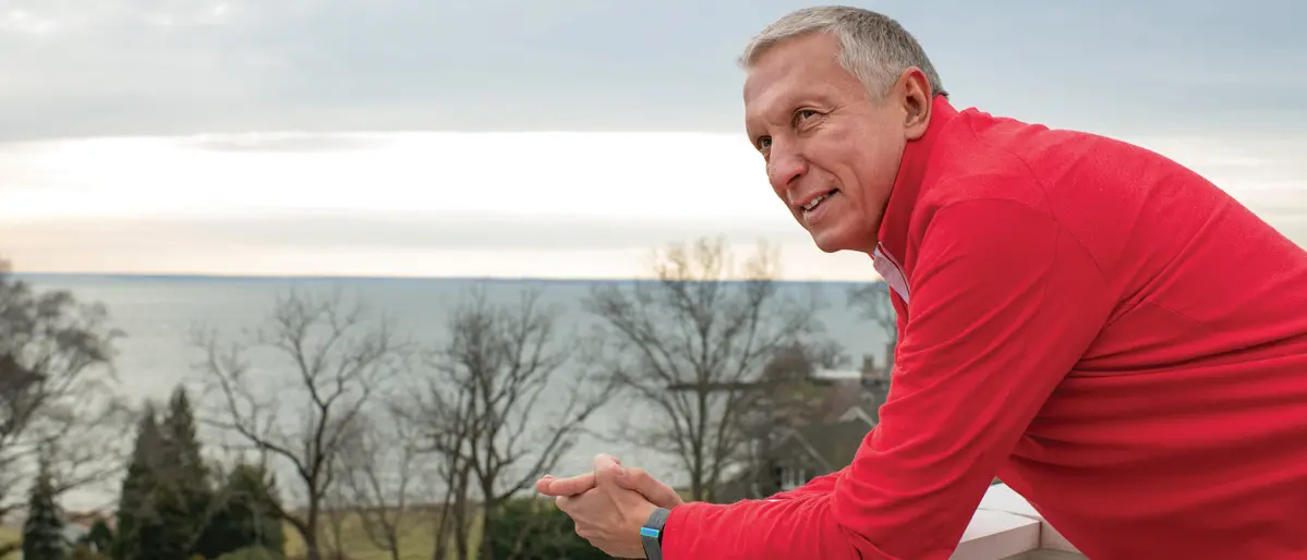 A white-haired man wearing a scarlet shirt leans on a railing on a roof-top porch with a view of the ocean. He has an interested, happy expression, as if he is looking, not at the pretty scene, but at someone talking next to him.