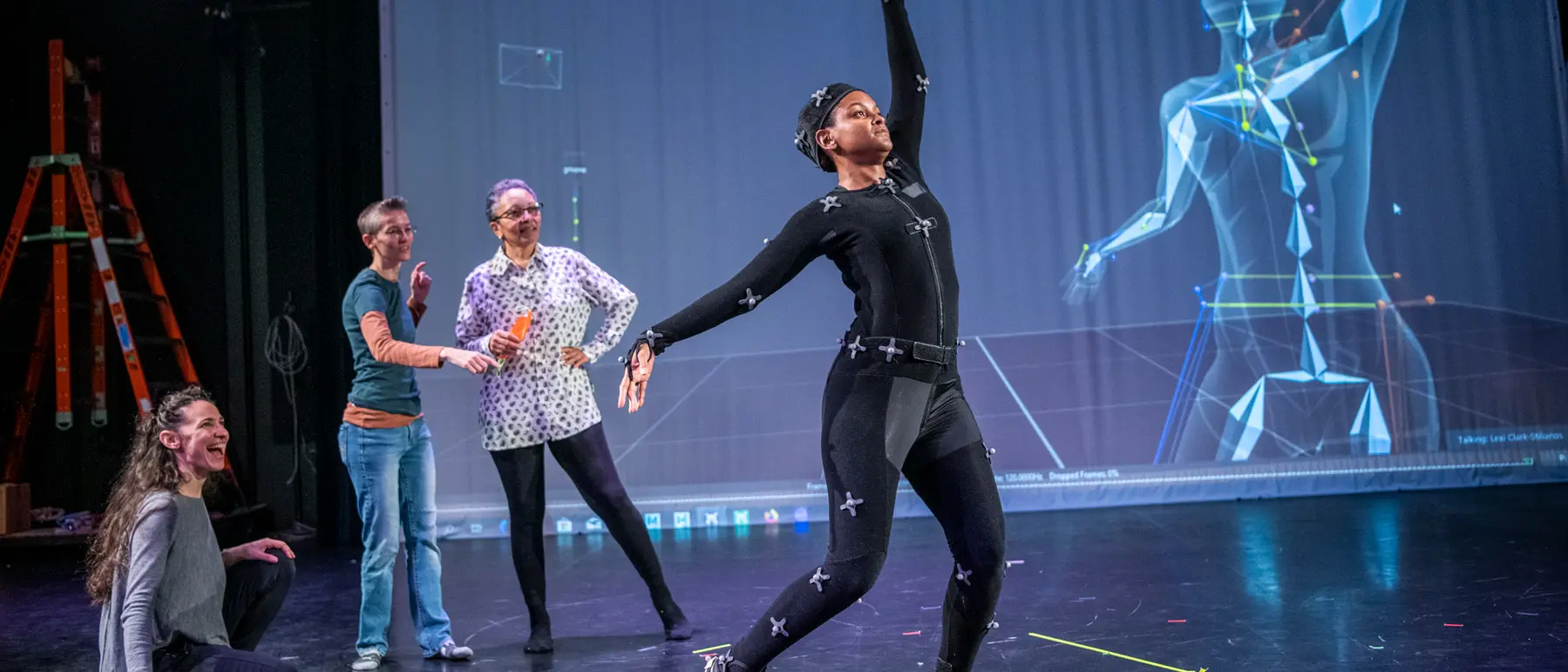 On a stage, a dancer in a black body suit and sensors steps forward and elegantly spreads her arms as three women behind her watch excitedly. A computer-generated geometrical illustration of the dancer highlights where her bones are as she moves.