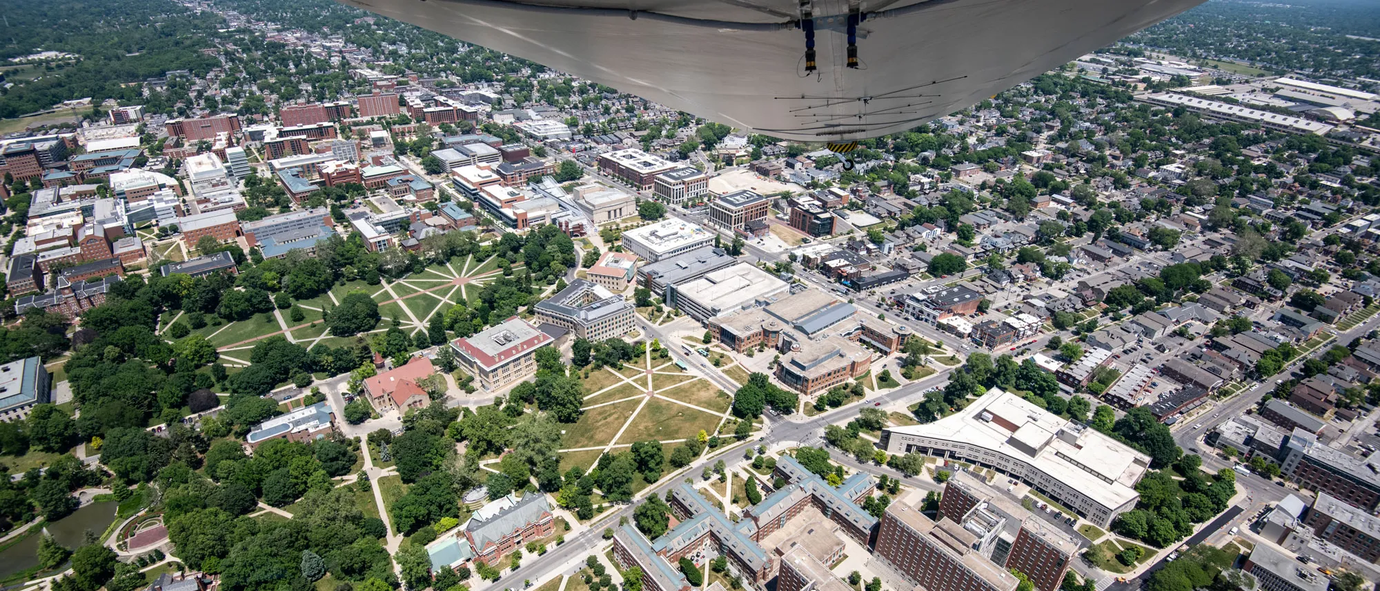 A photo taken from the blimp shows campus from high above. Prominent are the Oval, and the smaller similar field south of it, residence halls, the historical buildings on the Oval and new builds such as the music and theatre buildings. Trees look leafy and lush; sidewalks seem to glow; but some of the grass looks worn. The underside of the gondola can be seen at the top of the photo. 