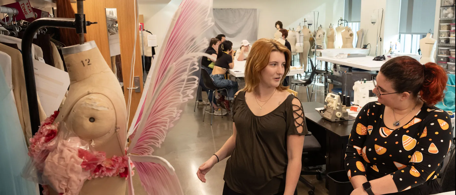 In the costume studio, a student a young woman with strawberry blond hair, smiles as she looks over her shoulder to chat with Catherine Huffman, the lead costume designer for She Kills Monsters, the play Emily is starring in.