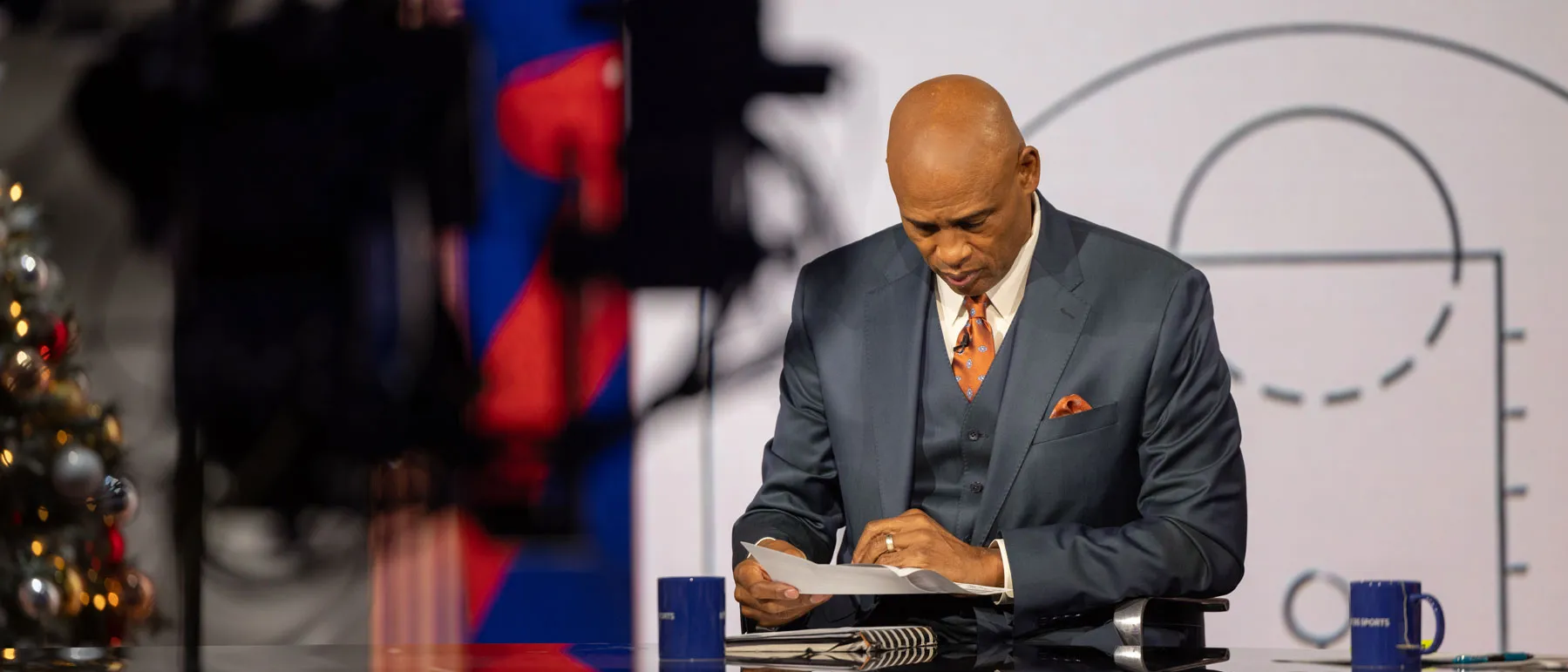 Kellogg sits at the studio broadcast desk, looking down and studying papers in he holds in both hands. A large camera is positioned in the foreground, and a sports themed graphic fills the background. 