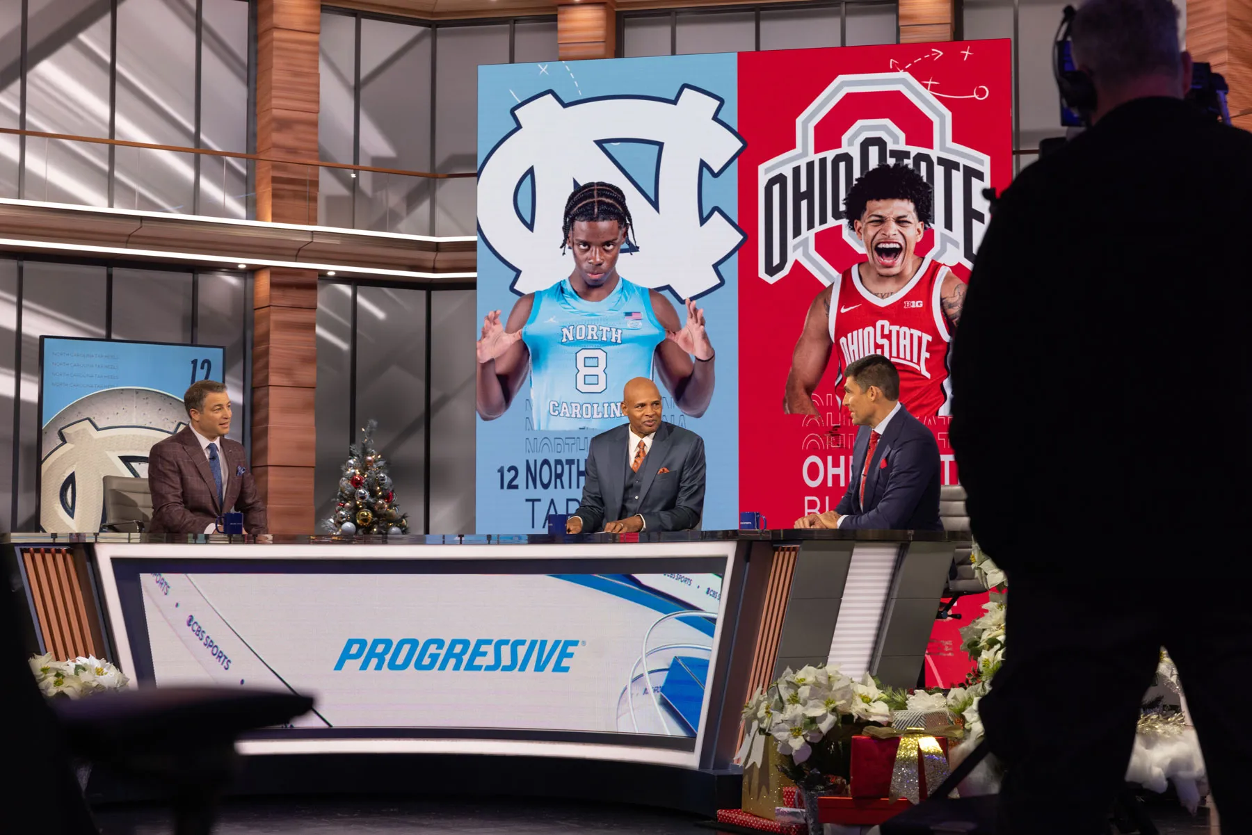 Three sports commentators, including Clark Kellogg, sit behind a studio desk during a televised sports segment, with large graphics of two basketball players displayed on screens behind them. One is Ohio State player John Mobley Jr. A camera operator stands in the foreground, and holiday decorations are visible around the set.
