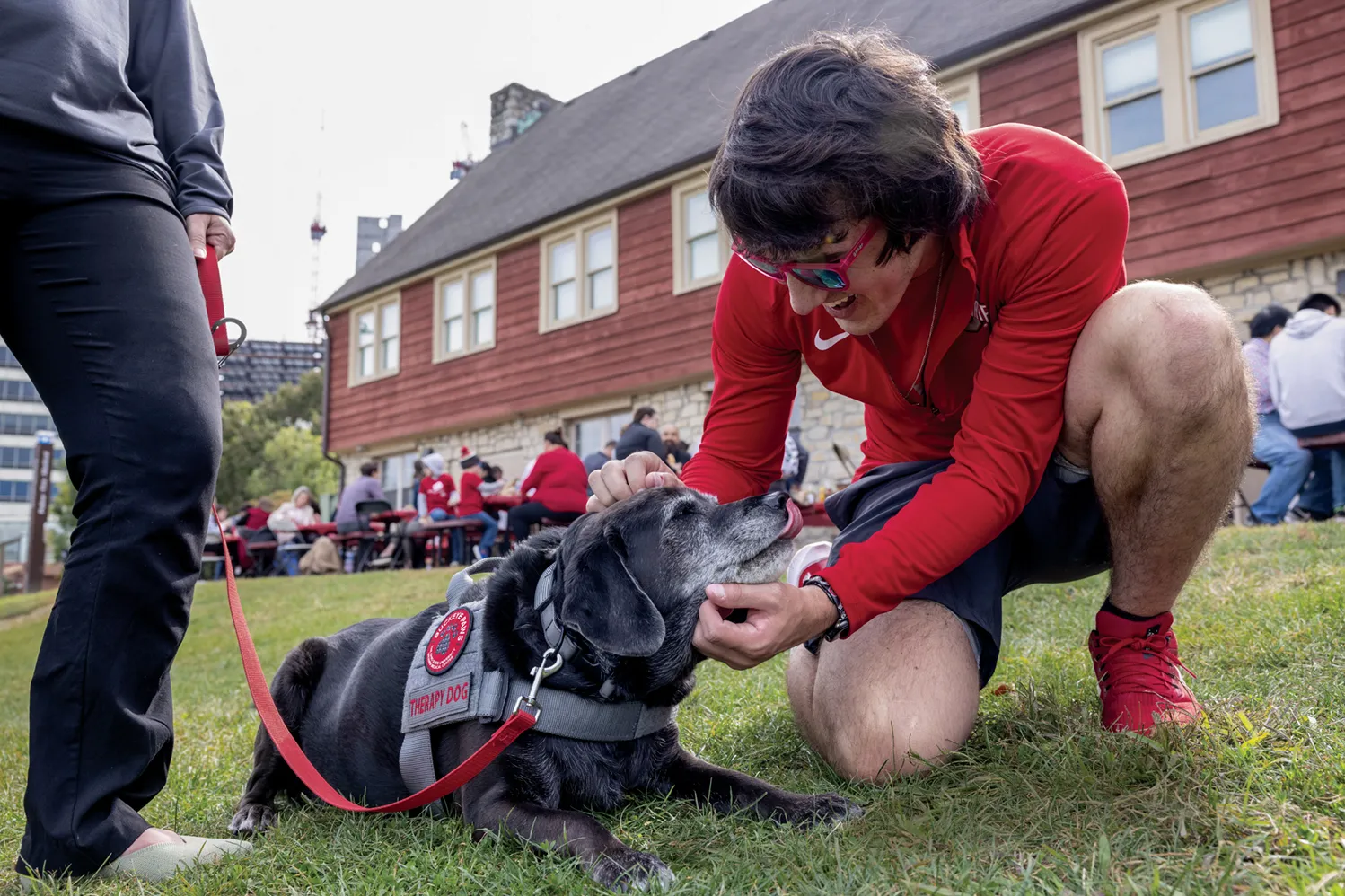 In the grass at an event, a young man wearing red sunglasses and an Ohio State shirt smiles as he kneels to pet a black dog. The dog seemingly gazes adoringly at him as it slightly licks its gray-speckled muzzle and wears a vest that says “therapy dog.”
