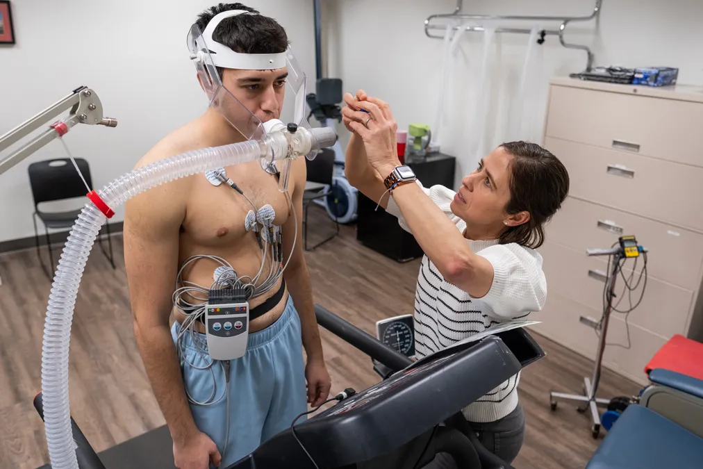 Carmen Swain reaches toward the head of a young male student standing on a treadmill. He wears head gear that supports tubing inserted into his mouth and connected to a machine off screen. He will breathe into the tubing while he runs.