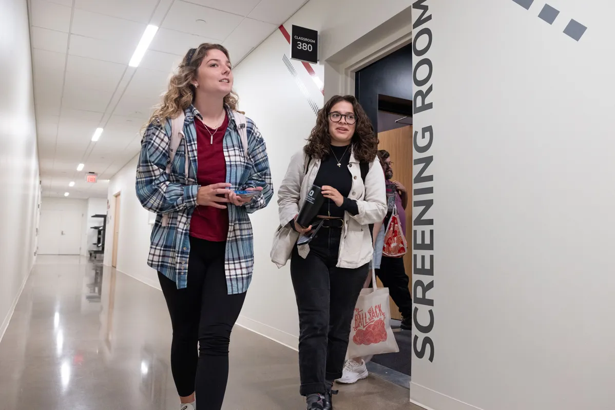 Two students, both young women, leave their classroom and begin walking up a hallway. The light paint on the walls and shiny floors make it apparent this is a new building. On the wall is painted the words “Screening Room.” The students both have wavy hair past their shoulders, wear button down shirts hanging open over more fitted shirts and black pants, and carry backpacks.