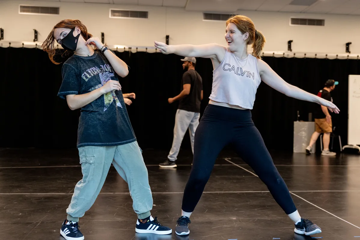 The student on the right, a young woman with her hair in a ponytail, laughs as she steps sideways and extends both arms, as if she has just swung at her class partner. That young woman, on the left, twists her head, making her hair swing, as if she has just been hit. They’re in a big, open classroom space.