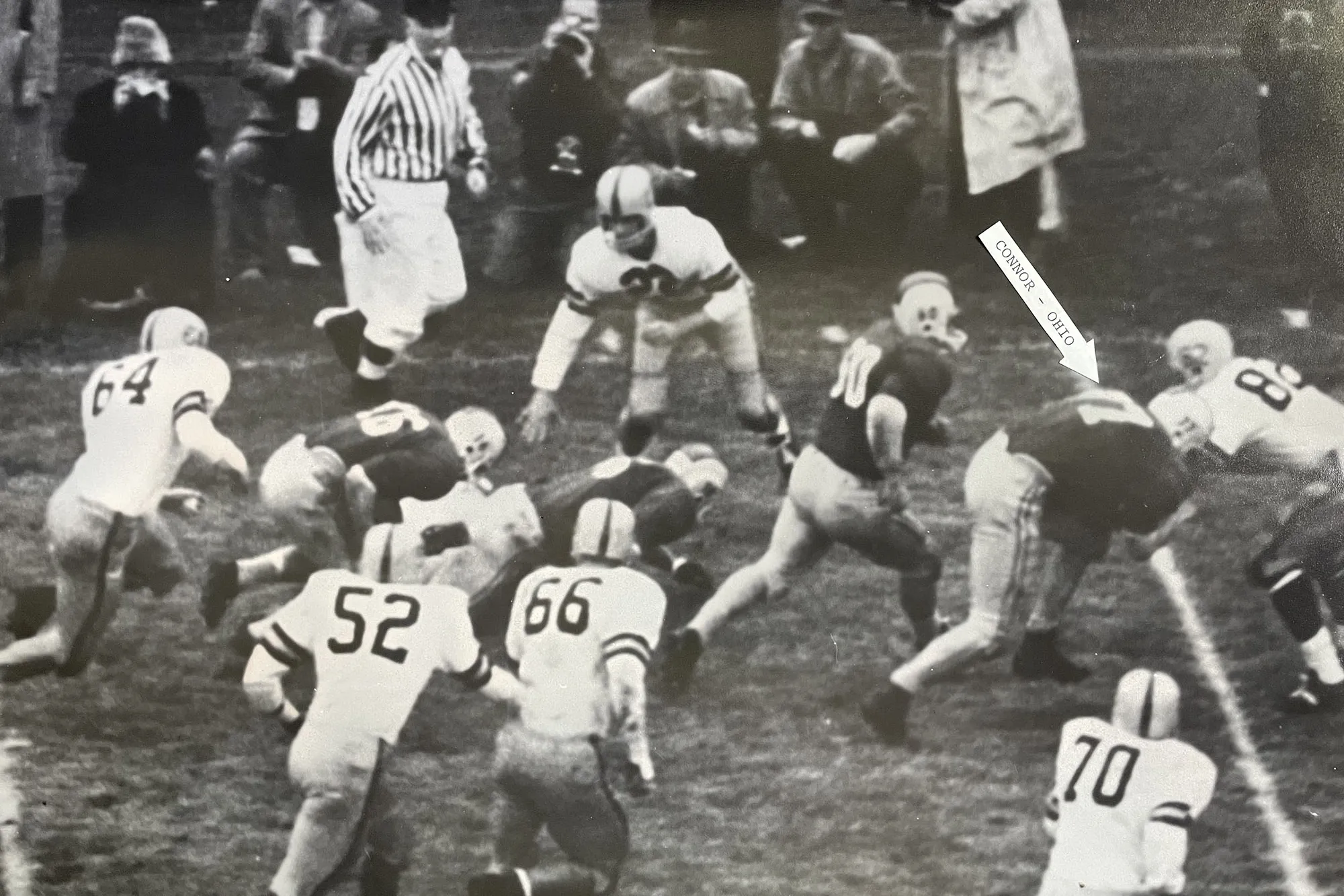 In a black-and-white photo from 1961, four Ohio State football players are surrounded by six defensemen from Iowa. The ball carrier is last in the line of Buckeyes while Daniel Connor is first, ramming an Iowa players.