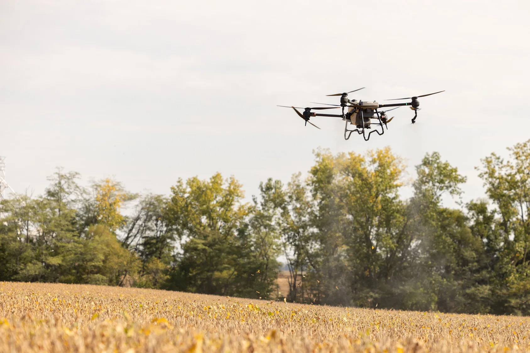 A drone flies above an out-of-focus field that is likely winter corn. In the distance there is a line of trees. The helicopter-like drone has three propellers pointing up, two sprayers on propeller arms pointing down, and thick wire stands hanging down from front and back. 
