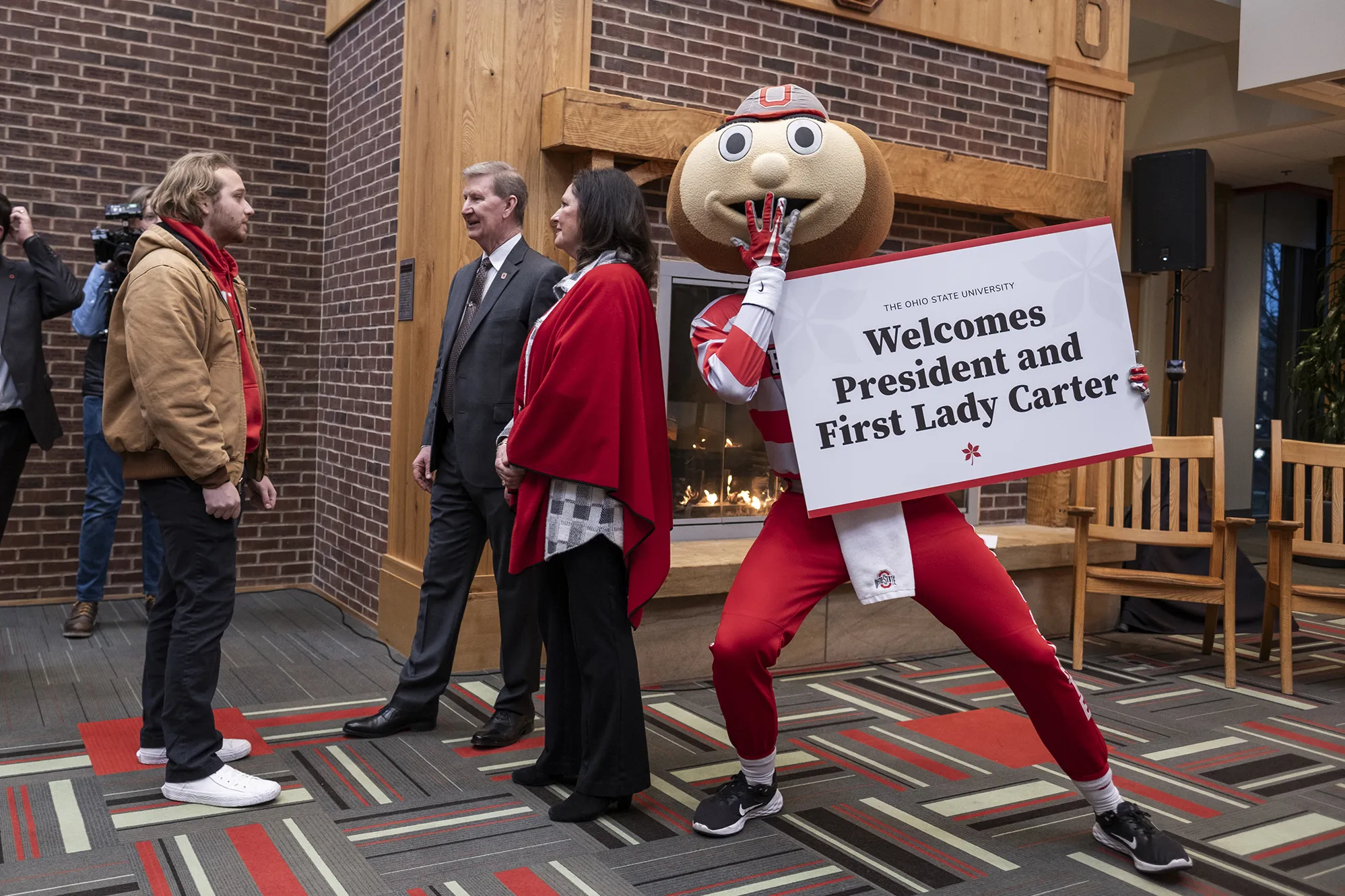 Brutus Buckeye holds a sign that says “Welcome President and First Lady Carter,” as the two speak with a student to Brutus’ right. Brutus is lunging sideways toward them, posing for the photo, and has his hand over his mouth as if laughing. (Brutus is a person wearing scarlet and gray and a giant buckeye as a head with a smiling face.) The Carters make eye contact with the student as they chat with him.
