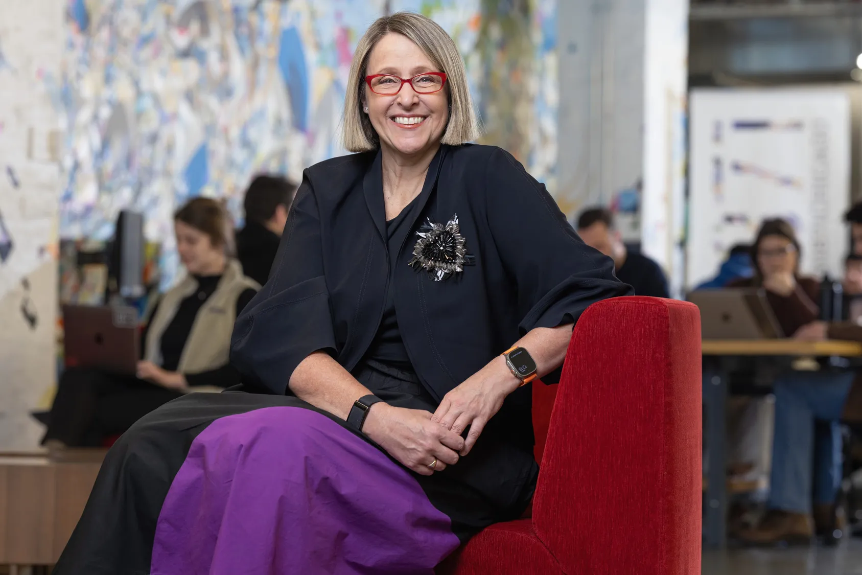 Betsy Ziegler sits on a modern upholstered chair in a space where many people in the background work or chat at small tables. A wall-size painting in the background adds to the lively feel of the scene, while Betsy poses with a genuine smile at the center of it. The middle-age white woman looks comfortable and stylish, with a chin-length haircut, stand-out glasses, modern-shaped clothes and a striking beaded broach that shines but also looks organically shaped.