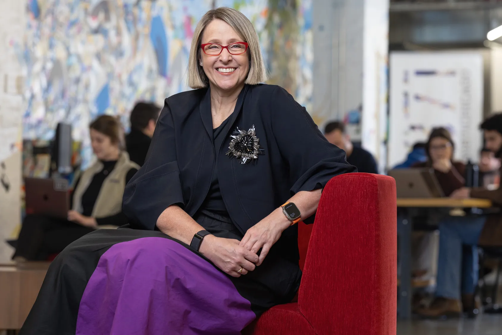 Betsy Ziegler sits on a modern upholstered chair in a space where many people in the background work or chat at small tables. A wall-size painting in the background adds to the lively feel of the scene, while Betsy poses with a genuine smile at the center of it. The middle-age white woman looks comfortable and stylish, with a chin-length haircut, stand-out glasses, modern-shaped clothes and a striking beaded broach that shines but also looks organically shaped. 