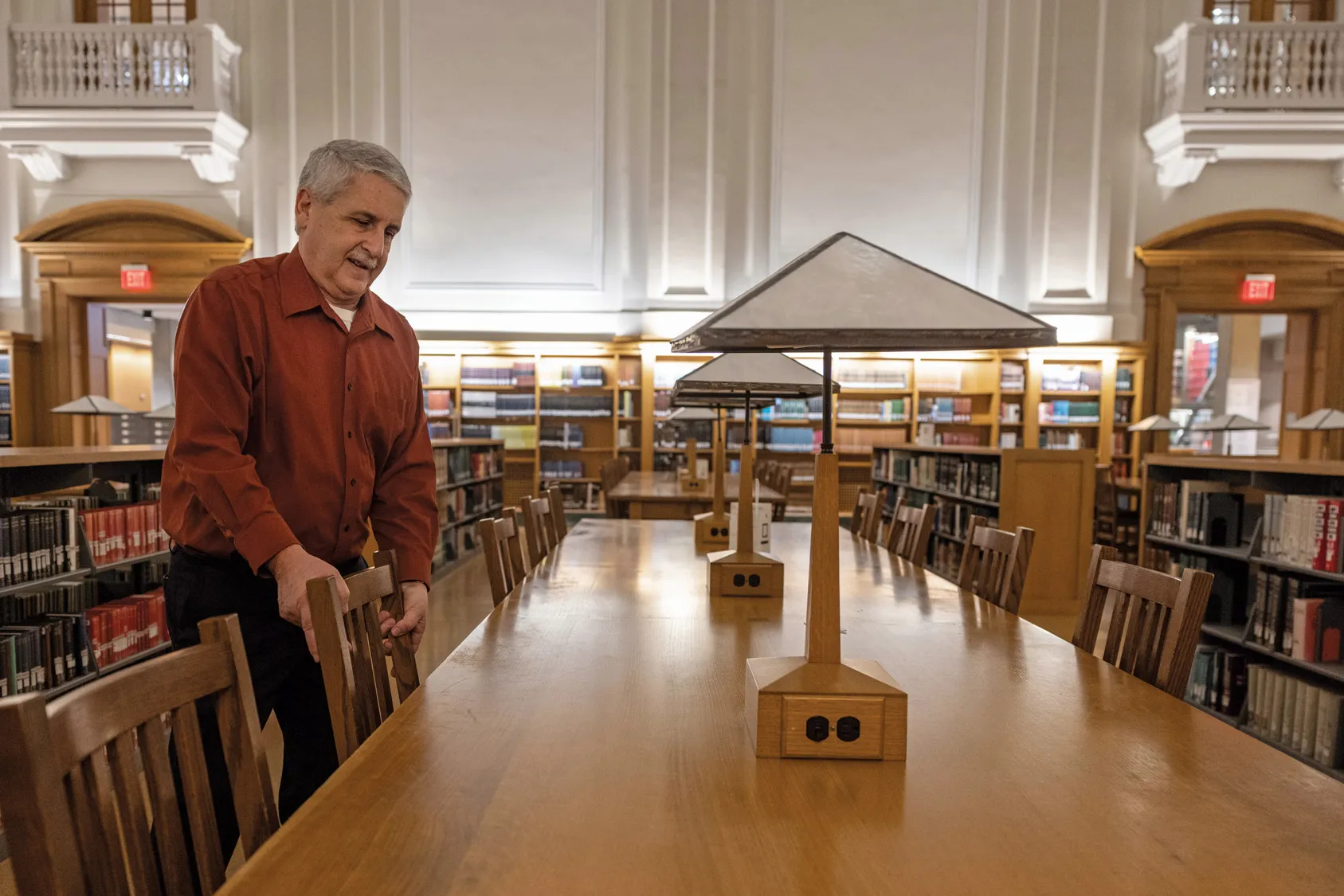 In a big hall, books line the distant wall and low shelves that run along a table as long as a handful of traditionally-sized dining room tables put together. The only person in the room is an older white man straightening one of the wooden, straight-backed chairs. Art-deco style lamps sit in the center of the table. 