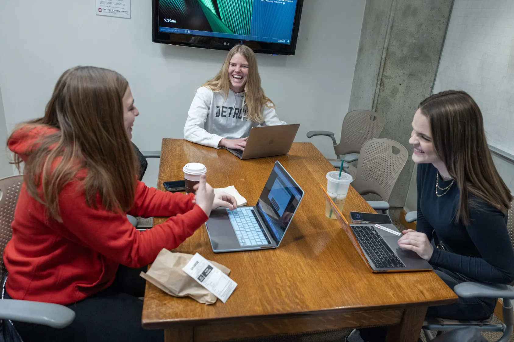 Inside a small meeting room with a screen on the wall, three young white women sit at a table, each with an open laptop and a grin, as they talk. They seem to be enjoying the work they are doing together. 