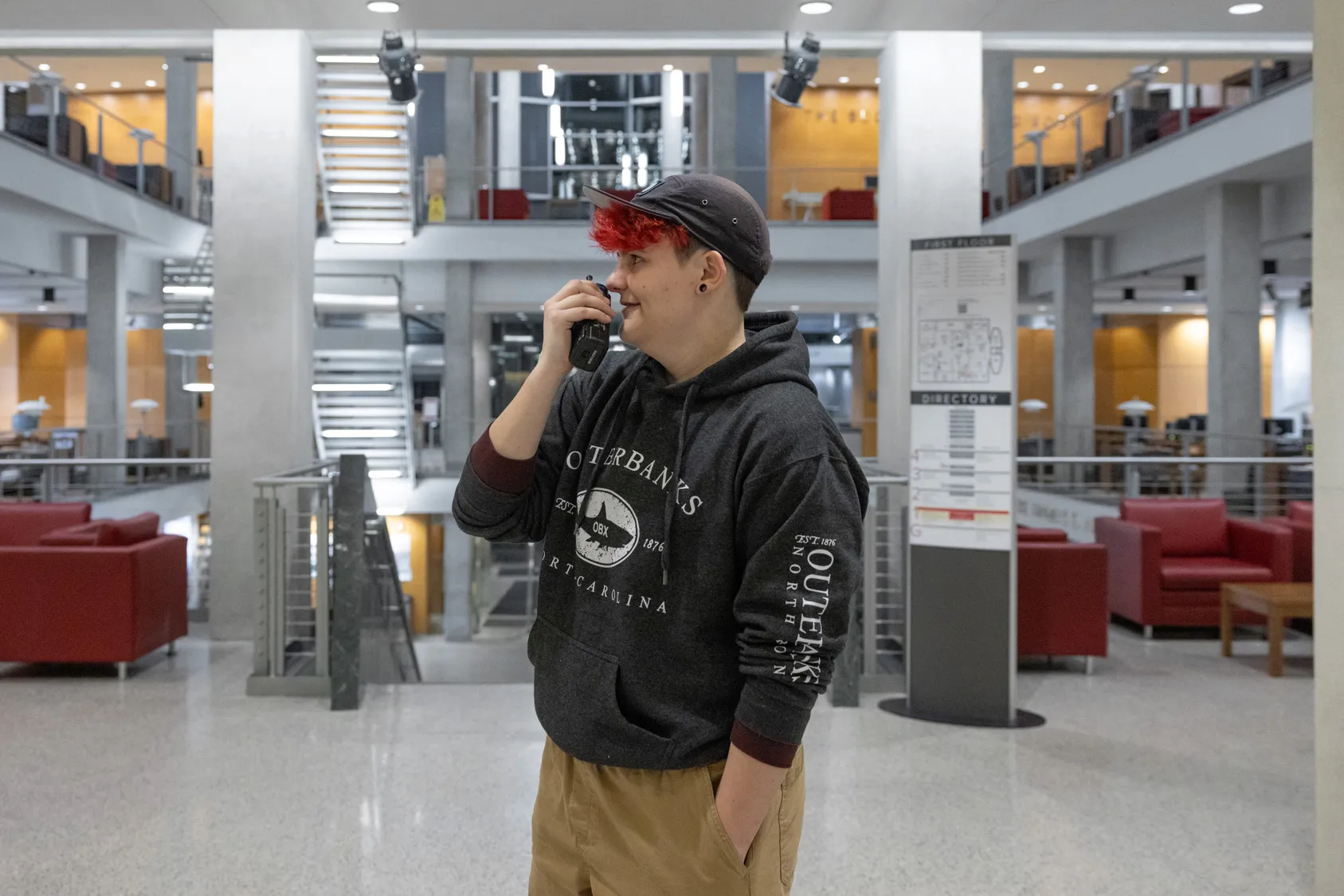 A white student in a sweatshirt, khakis and ballcap over red-orange-dyed hair speaks into a walkie-talkie while on a lower floor of the library. The lights are all on, but no one else can be seen. 