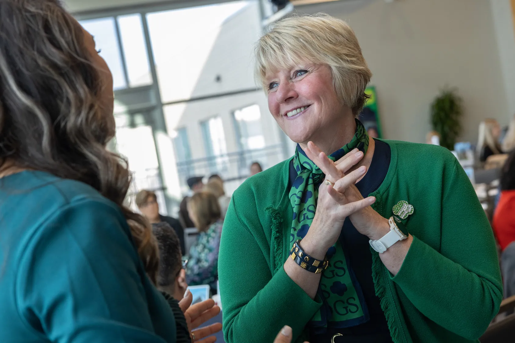 From this photo, you can tell that Tammy Wharton, a white woman with short blond hair and a pretty smile, has excellent listening habits. She seems super-engaged and even encouraging as she looks directly at the woman who’s speaking with her, who is pictured from behind. Other people are in the background of the photo, so you can see it’s a meeting or convention at a place where huge windows let in lots of natural light. 