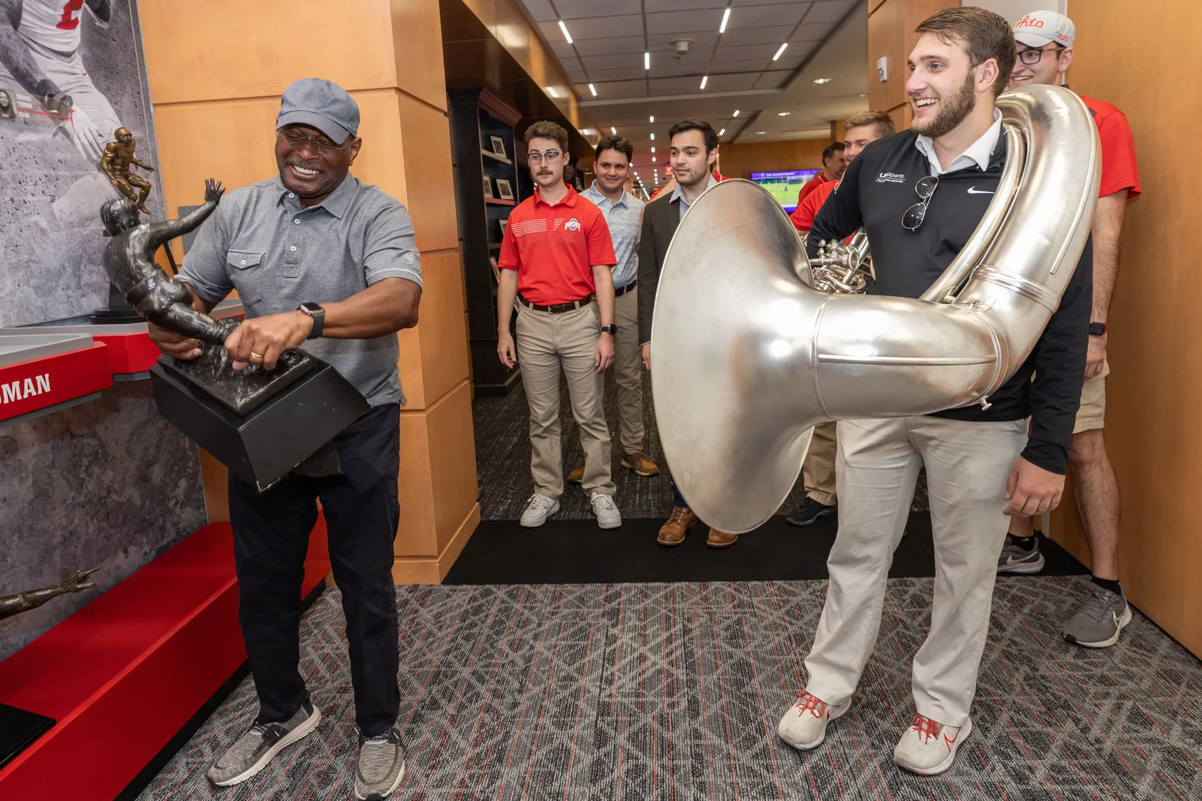 Archie Griffin hefts a replica of his Heisman Trophies—it must be heavy because you can see the muscles in his forearms. Watching are members of the marching band. The student in the front, a young white man with a beard, has a sousaphone wrapped around him. This was the day they invited Griffin to dot the i at a game this season. 