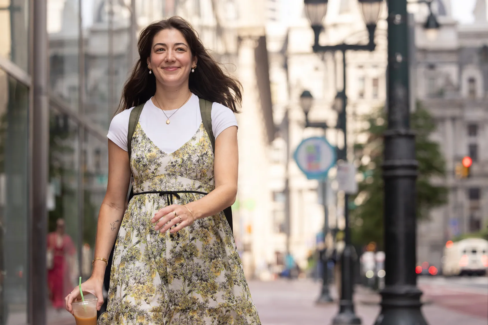 Tovah Kaiser smiles as she strides down a Philadelphia sidewalk. She’s a young white woman with long hair and a tattoo of a simplified fish on the inside of her right arm. She’s wearing a pretty and floaty floral dress over a plain white T-shirt along with gold earrings, necklace, bracelet and rings.