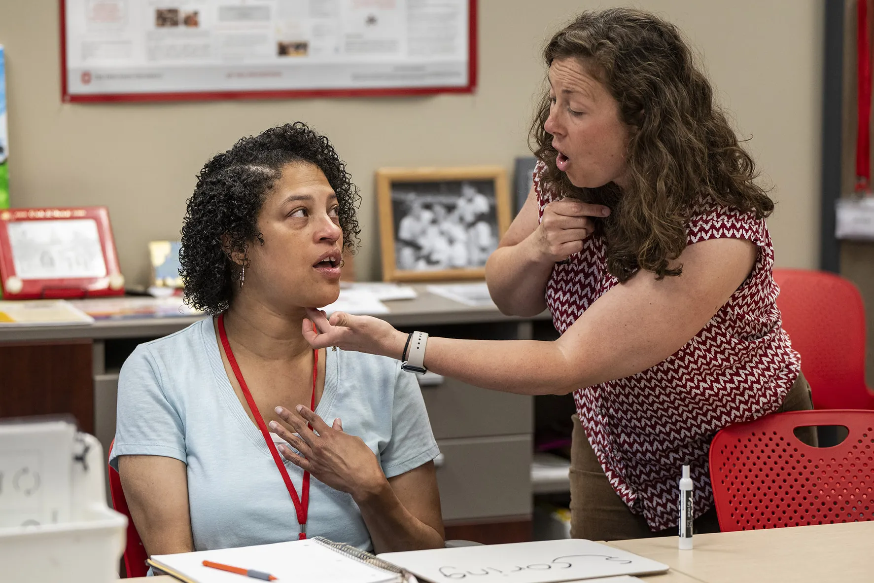 Amber Poindexter, sitting at a table, touches her chest as she watches the mouth of her speech therapist, Arin Sheeler. Arin is a curly-haired white woman leaning toward Amber, pointing at her own throat and touching the underside of Amber’s chin. They’re very focused on each other.