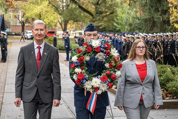 President Carter participates in a veterans ceremony with two students. Lines of ROTC students in dress uniform stand in the background.