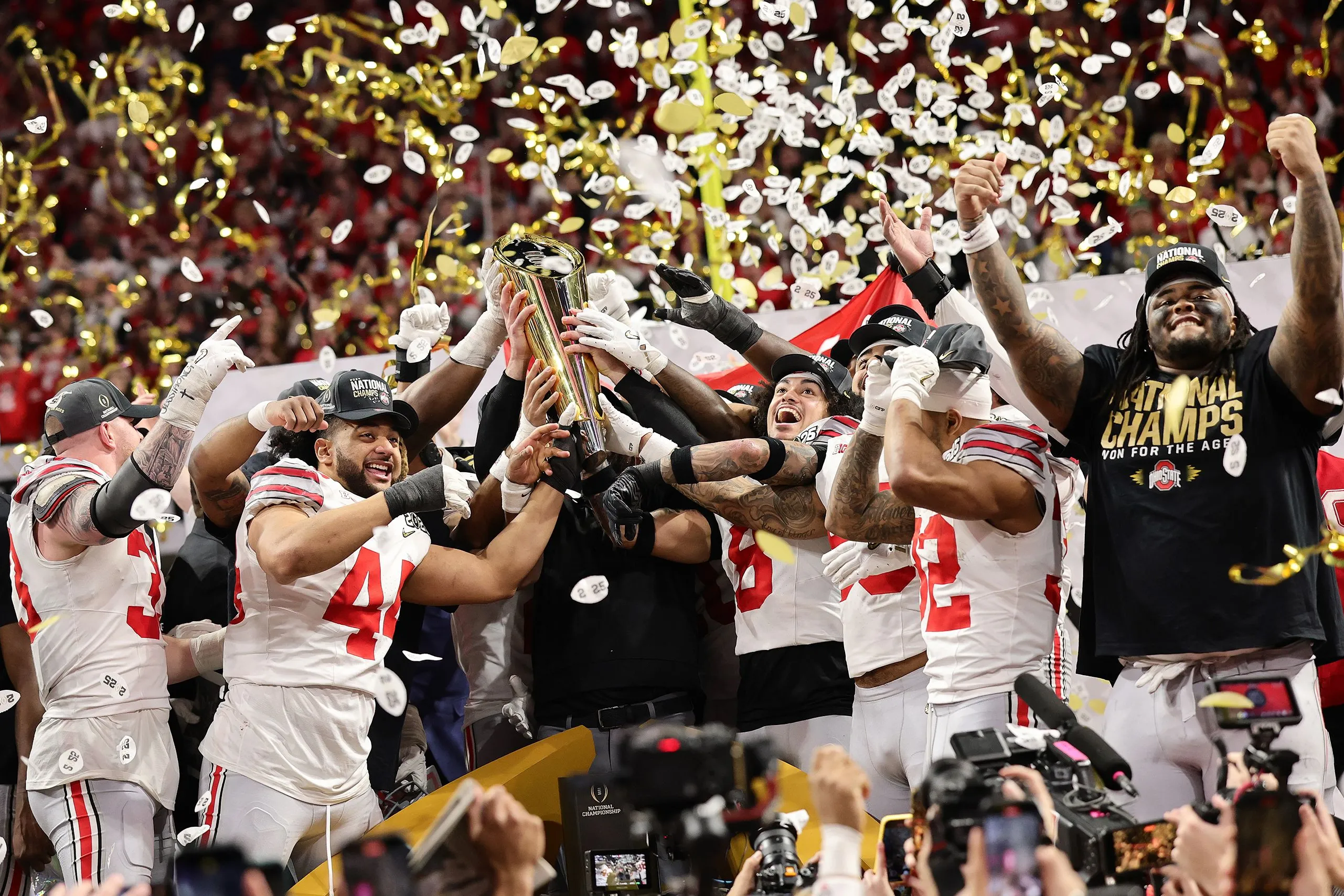 Buckeye football players cheer as they hoist up the national championship trophy. Confetti falls and they're amidst a huge crowd.