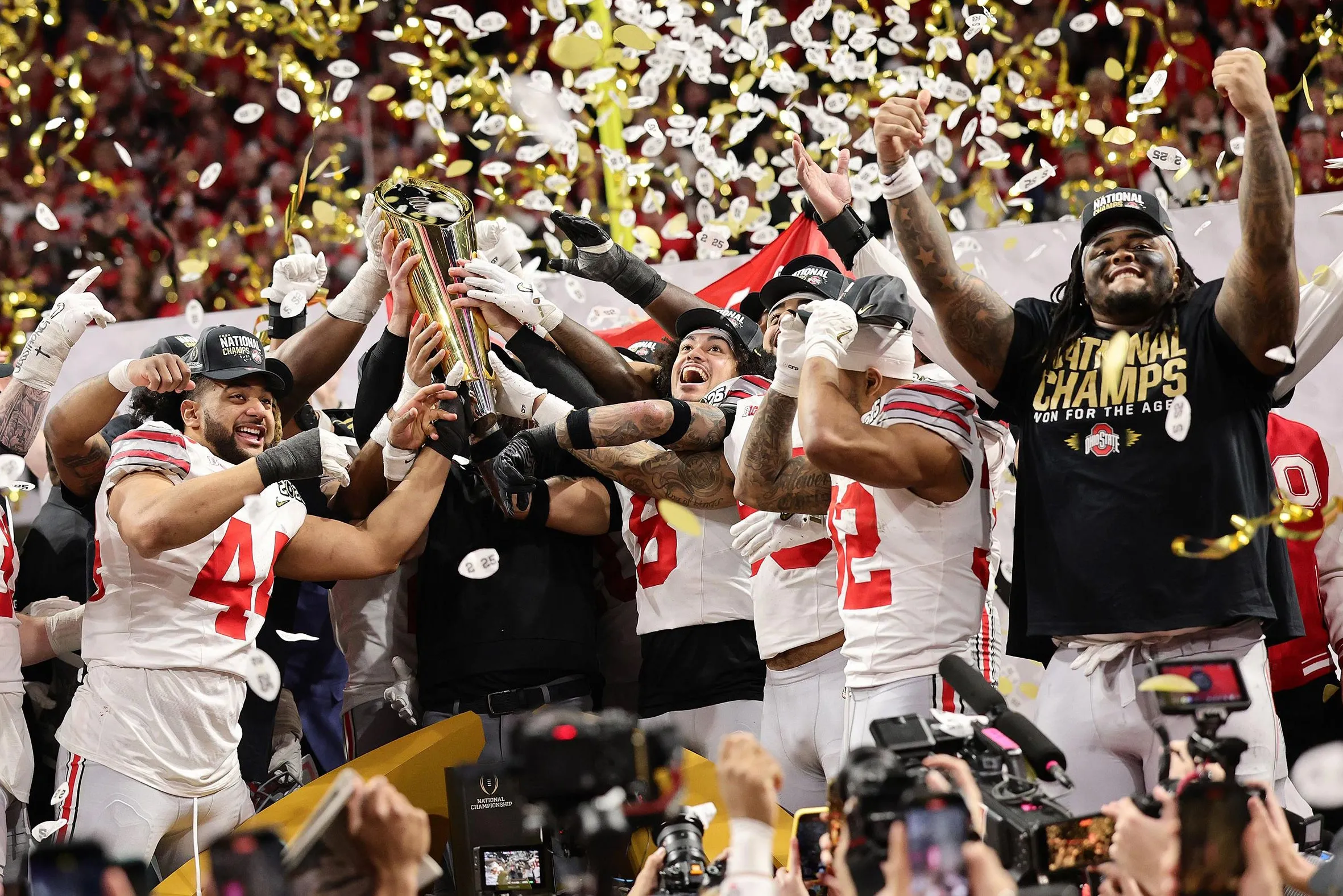 Buckeye football players cheer as they hoist up the national championship trophy. Confetti falls and they're amidst a huge crowd.