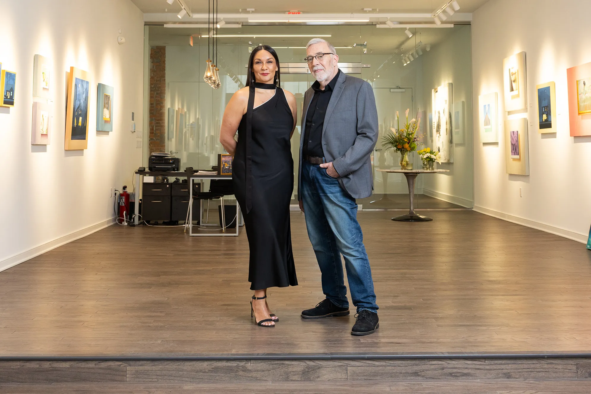 Michelle Brandt wears a black dress and Duff Lindsay wears jeans and a sports coat in a new art gallery. They seem elegant and thoughtful as they pose standing next to each other and slightly smiling.