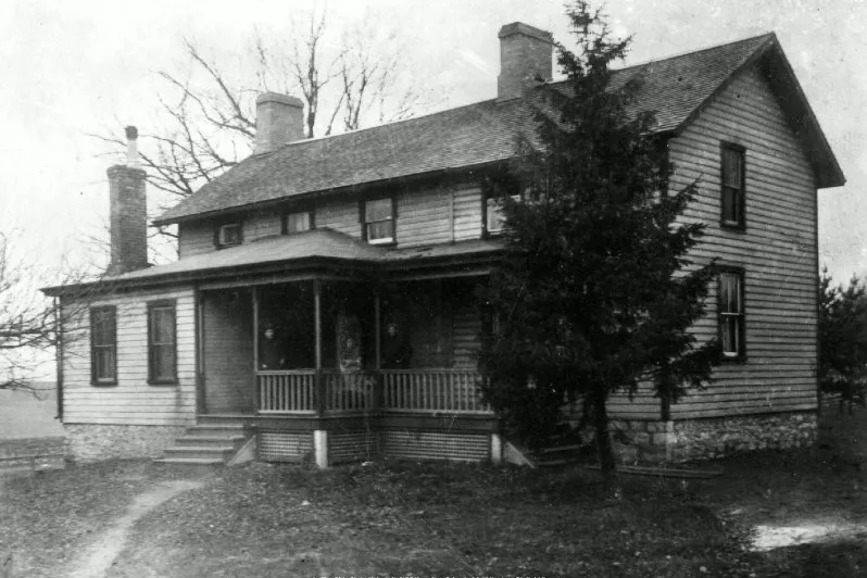A black and white photo shows an old two-story wooden farmhouse. 