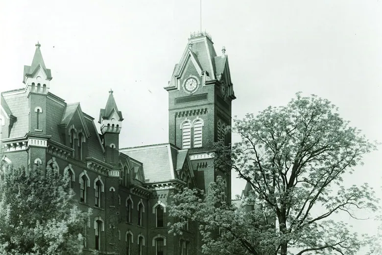 A black and white photo shows the original University Hall. The the belltower is taller than a full-grown tree in front of the building.