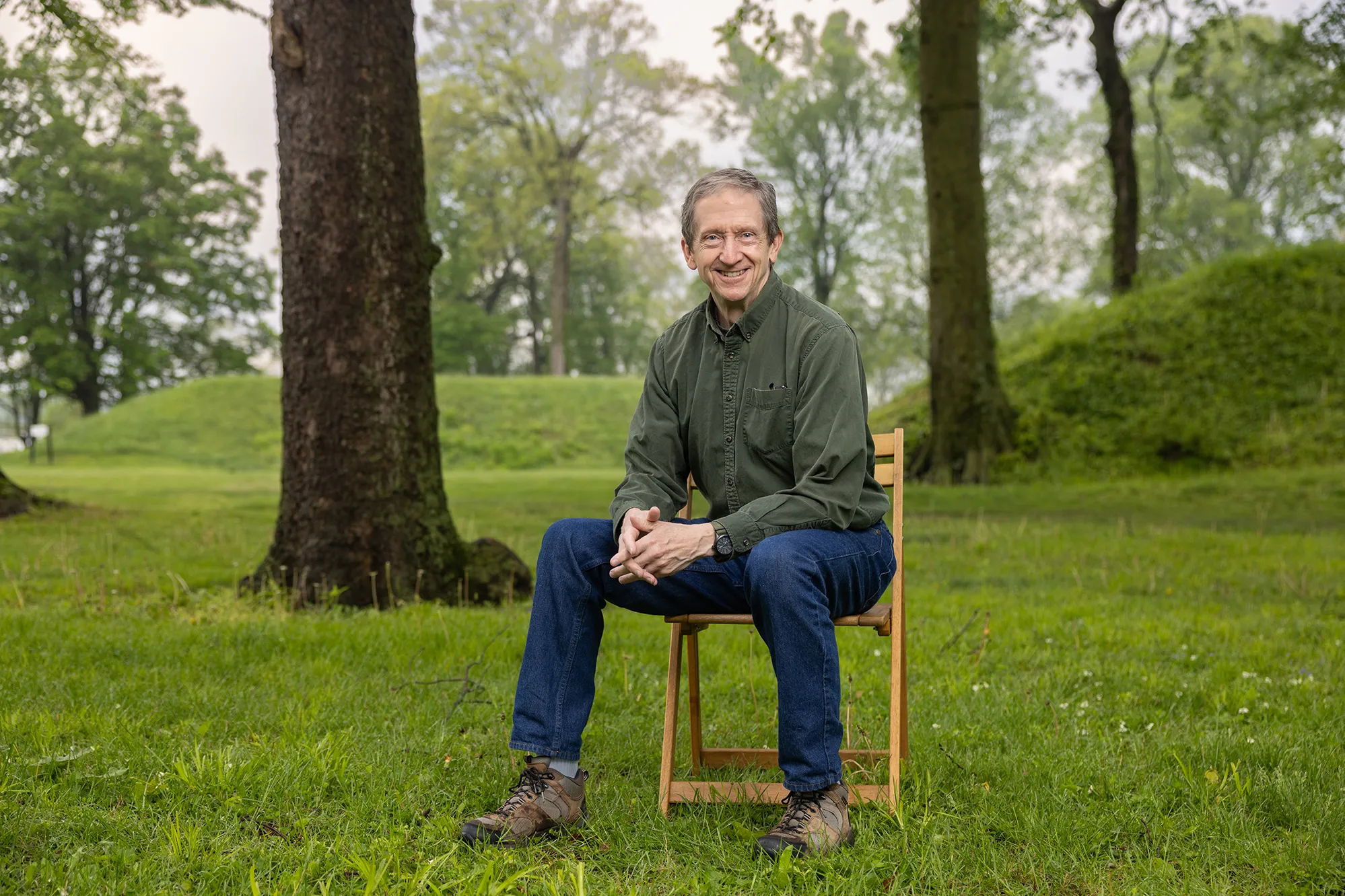 Bradley Lepper, a gray-haired white man in jeans and an earthy colored button down, sits on a chair at the Great Circle Earthworks. Around him are trees and behind him, grass-covered mounds rise. The portions of the mounds in the photo look to be about as tall as an adult, or at least shoulder height.