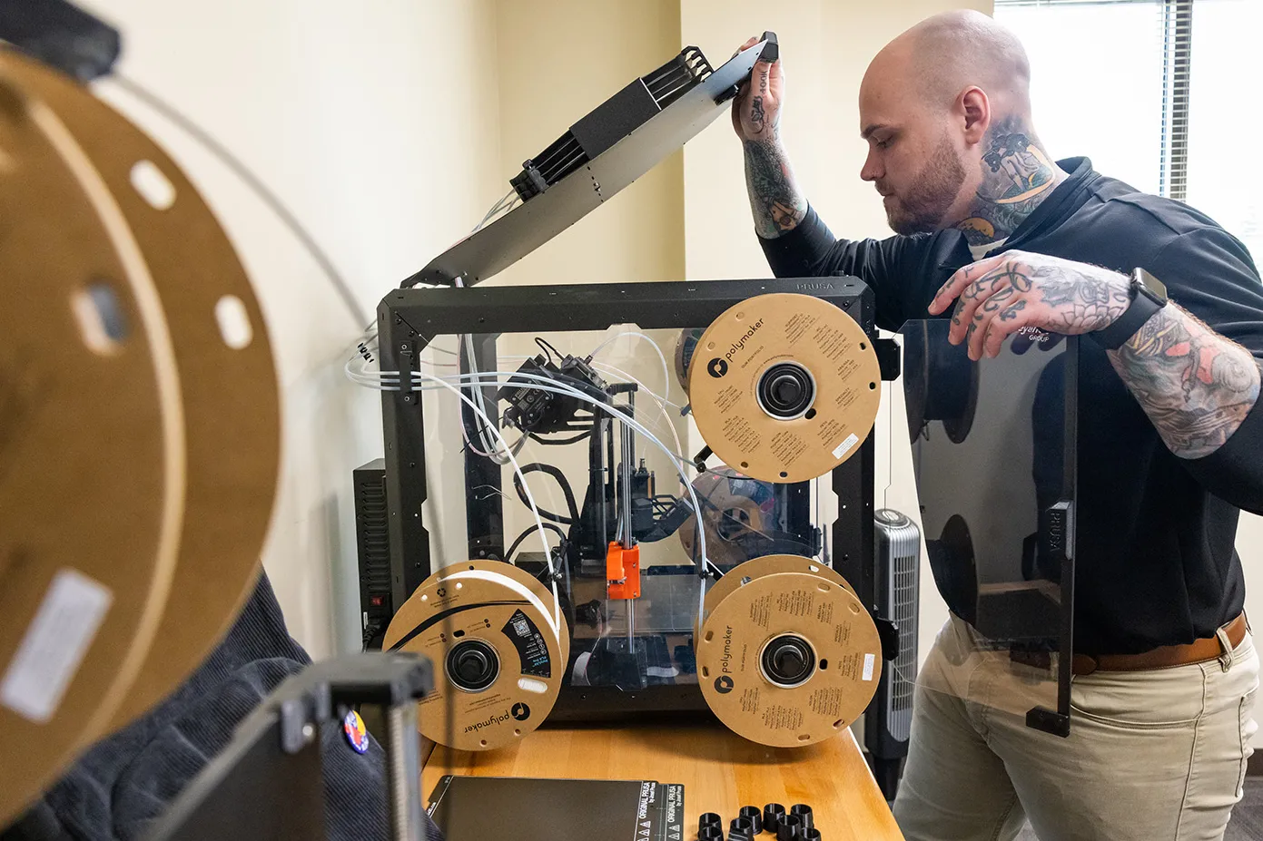 Zack opens the top of a 3D printer to check a component. The printer is a clear box with three spools of plastic mounted on the side and the printing hardware looking futuristic inside the box.