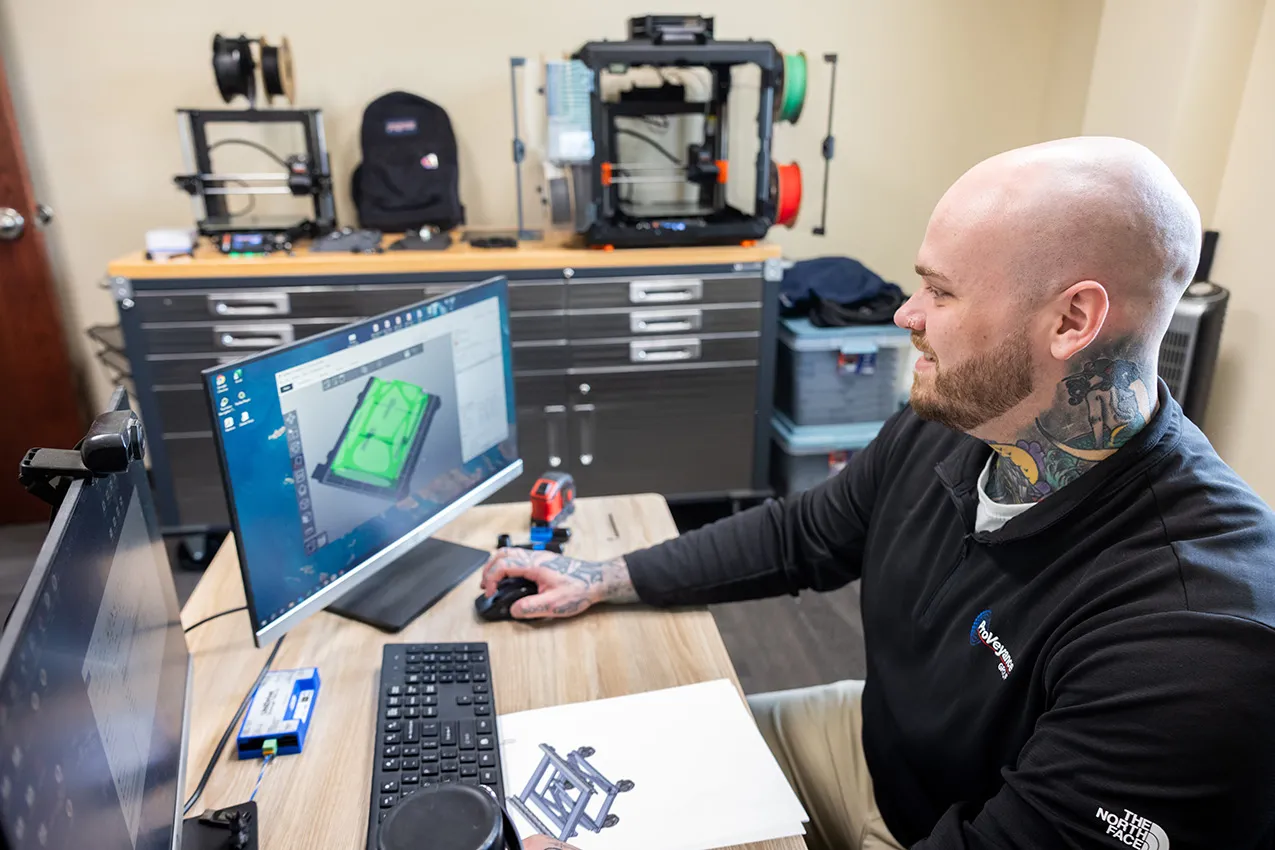In his office, Zack works on a computer. The screen shows a piece he's designing. 3D printers stand on a 6-foot toolbox along the far wall. Zack's smiling--it appears he enjoys his work.